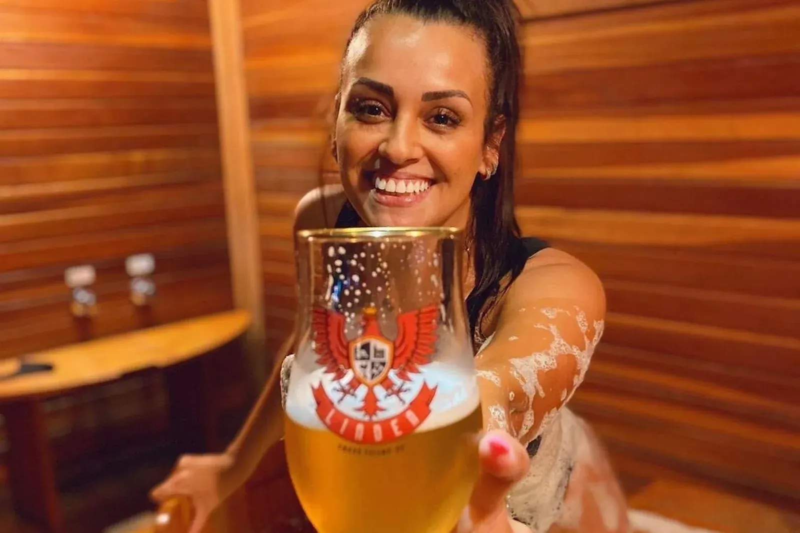 Uma moça no ofurô estendendo um copo de chope com o brasão da LindenBier