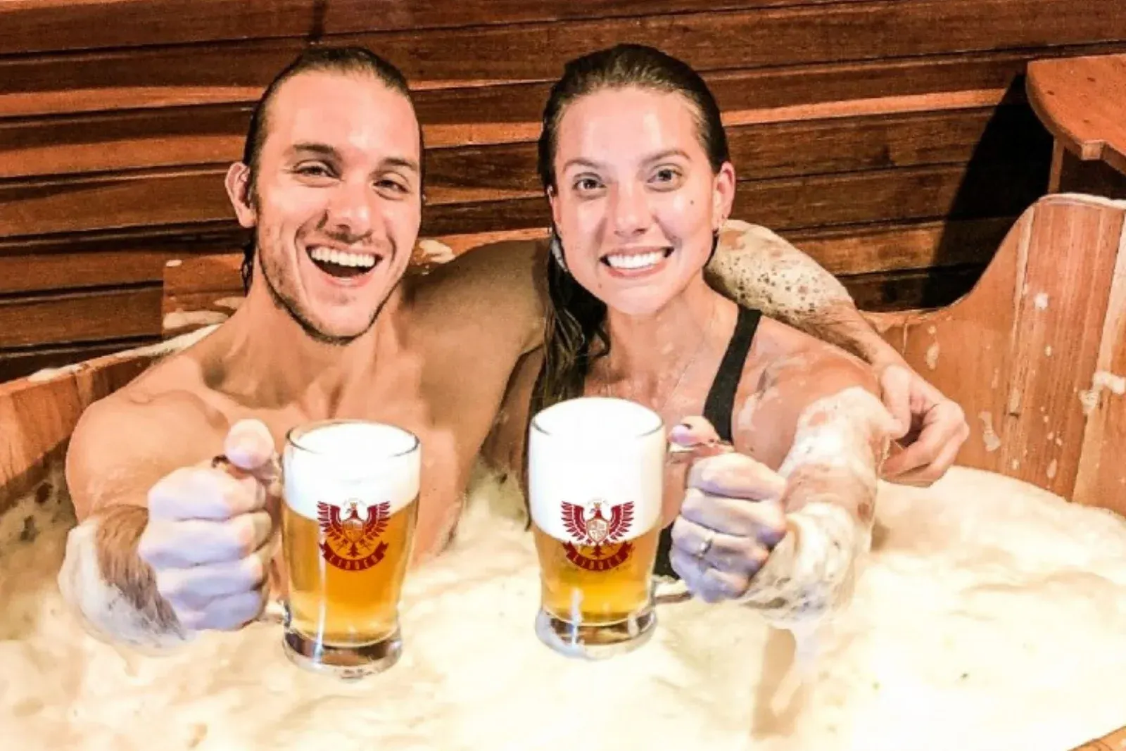 Um casal sorridente num ofurô de madeira com espuma, brindando com canecas de chope LindenBier