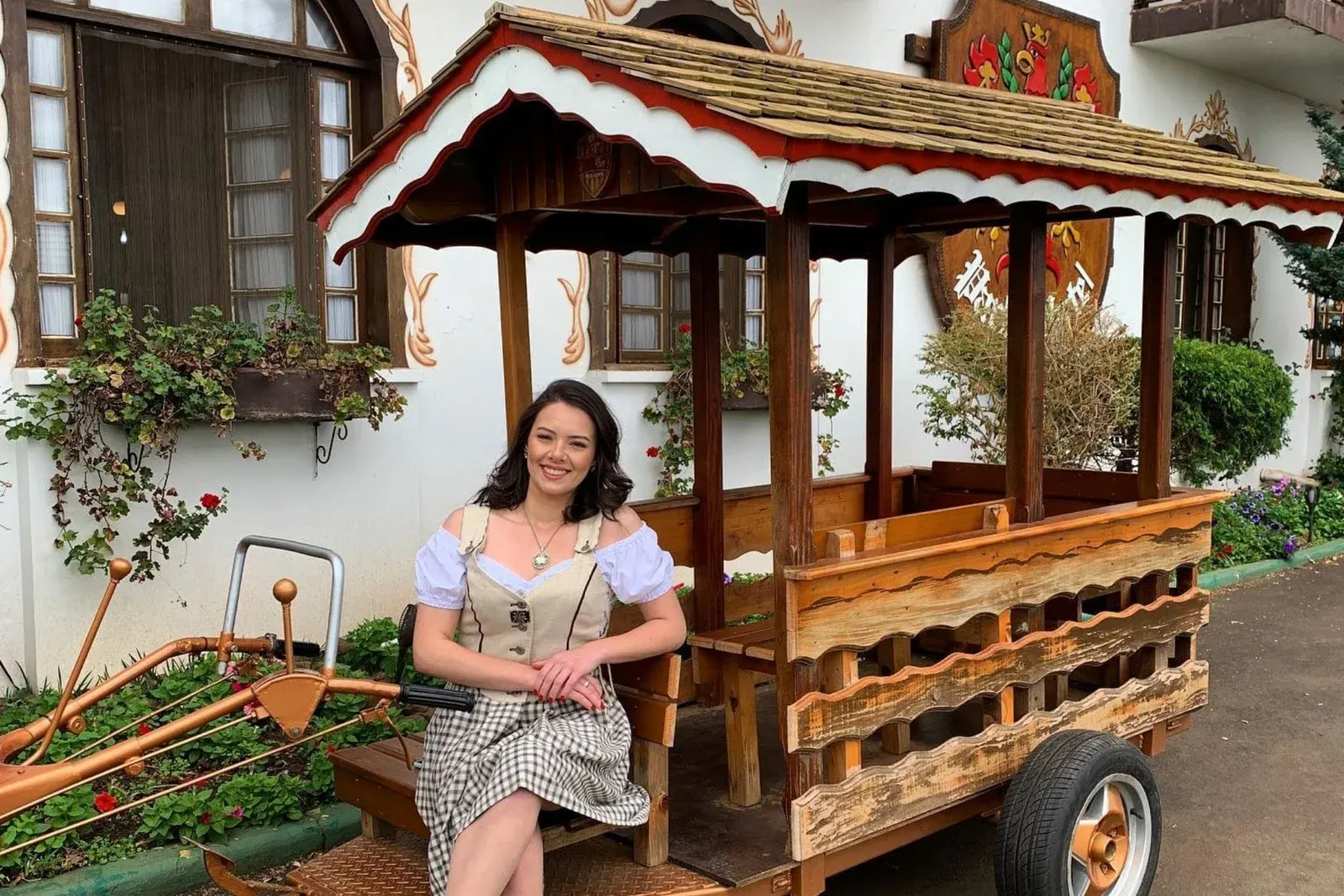 A Bierwagen, uma carroça de madeira entalhada, com uma moça de dirndl sentada ao lado, diante da fachada tirolesa do hotel